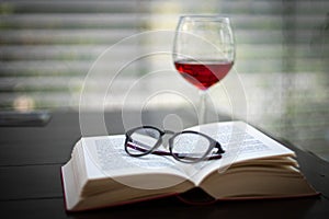 Pair of glasses lies on an open book and in the background is a wine glass