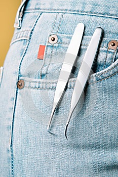 Pair of high-quality tweezers, using for isolating and gluing lashes, hanging from pocket of blue denim jeans of woman.
