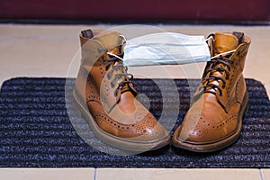Pair of Mens Leather Tanned Brogue High Boots Covered With Facial Medical Mask on Door Mat as a Virus Protection Sign