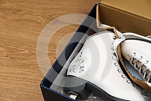 A pair of new white figure skates for women in the opened box. The box lies on a light brown wooden background. Selective focus.