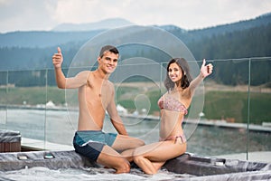 Pair with perfect figure sitting on the Jacuzzi lowered feet in the water and showing thumbs up gesture of good class