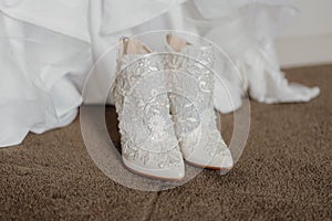 Pair of pristine white wedding shoes on a brown carpet floor with a wedding dress in the background