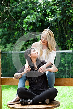 A pair of proud lesbian in outdoors sitting on a wooden table, blonde woman is hugging a brunette woman, in a garden