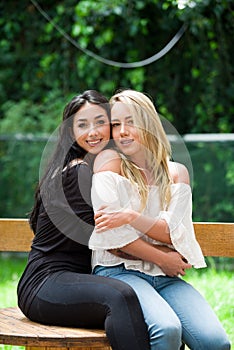 A pair of proud lesbian in outdoors sitting on a wooden table, brunette woman is hugging a blonde woman, in a garden