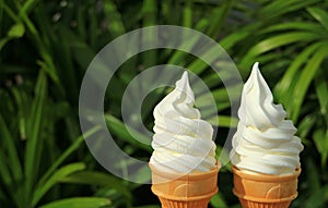 Pair of pure white milk soft serve ice cream cones in the sunlight, with blurred green foliage