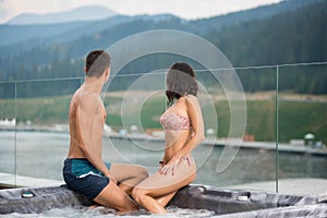 Pair sitting on the Jacuzzi lowered feet in the water and look at the scenery of nature turned away from the camera