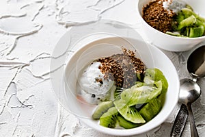 A pair of small dessert bowls with fresh kiwi fruit, white ice cream, yogurt and chocolate chips on a white stone background