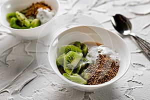 A pair of small dessert bowls with fresh kiwi fruit, white ice cream, yogurt and chocolate chips on a white stone background