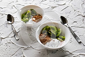 A pair of small dessert bowls with fresh kiwi fruit, white ice cream, yogurt and chocolate chips on a white stone background