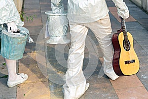 Pair of street artist painted in white silver paint walking in city park. Living statue performer with guitar