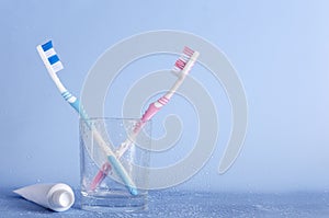 Pair of toothbrush in the glass cup, tube with toothpaste on the blue wet surface