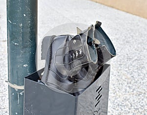 a pair of used black boots made in black leather abandoned in a black bin. Boots and bin at a post in the sidewalk of the street.