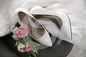 Pair of white high heel shoes, flowers and wedding dress on chair