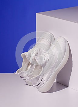 Pair of white sneakers stand on white geometric podium on blue background.