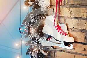 Pair of white vintage leather skates with red laces hanging on old rustic brick wall with garland lights on christmas tree