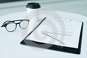 A pair of women`s glasses and a pen lying on top of a white sheet of paper on wooden surface. The sheet has a coffee stain. Copy