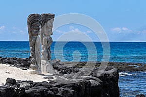 Pair of wooden status Tikis, place of refuge Honaunau, Hawaii. White sand, wall of black lava rock, ocean and blue sky.