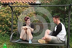 A pair of young people talking on a garden swing.