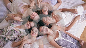 Pajama party, six smiling young women in stylish sleepwears lie on the bed and look directly into camera in room
