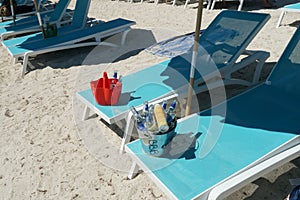 Pale blue chairs on the sandy beach on top of which are red plastic and metal buckets full of empty beer bottles.
