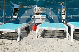 Pale blue chairs and umbrellas on the sandy beach next to which is a red plastic bucket full of empty beer bottles.