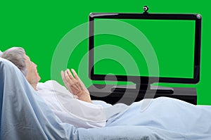 Pale old woman lying in bed waving to a chroma key big monitor screen.