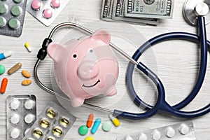 Pale pink ceramic piggy bank, stethoscope, money and pills on white wooden table, flat lay. Medical insurance