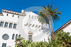 A palm tree and a white Spanish architecture style building with ornate windows and trim under a beautiful blue sky