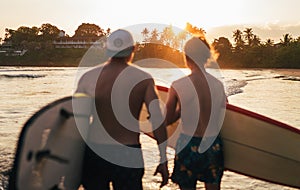 Palm trees lightened with the sunset sun. with father with teenage son walking with surfboards by the sandy ocean Dewata beach in