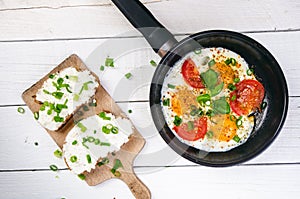 Pan of fried eggs with tomatoes, cheese, spring onion, herbs on a white table. Bread with spread. White wooden table. Concept of f