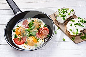 Pan of fried eggs with tomatoes, cheese, spring onion, herbs on a white table. Bread with spread. White wooden table. Concept of f