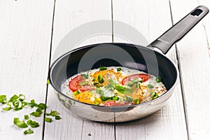 Pan of fried eggs with tomatoes, cheese, spring onion, herbs on a white table. White wooden table. Concept of food. Breakfast time