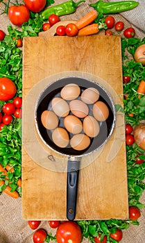 Pan full of eggs on a wooden surface surrounded by vegetables - green salad, tomatoes, cherry tomatoes, carrots and pepperoni