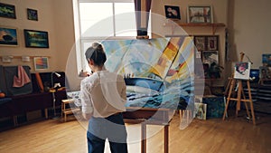 Pan shot of serious girl professional painter working in studio painting marine landscape with tempera paints holding