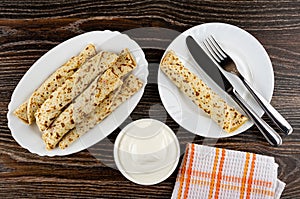 Pancake rolls in dish, bowl with sour cream, pancake roll with filling, knife and fork in plate, napkin on wooden table. Top view