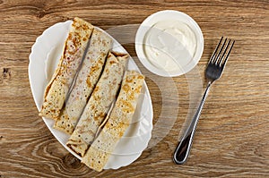 Pancake rolls in white plate, bowl with sour cream, fork on wooden table. Top view