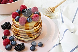 Pancakes with berries and honey on a pink plate over white wooden background, side view.