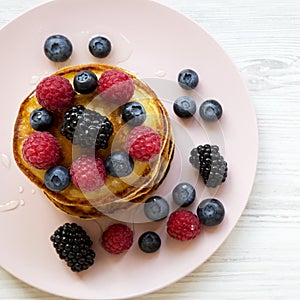 Pancakes with berries and honey on a pink plate on white wooden backfround, overhead view.