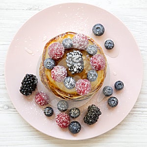 Pancakes with berries, honey and sugar on a pink plate on a white wooden table, overhead view. Flat lay, top view, from above. Clo