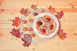 Pancakes with homemade plum  jam on white plate, fresh plums and autumn maple leaves on wooden background.  Toned image