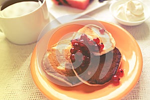 Pancakes in orange plate and glass of sourcream with red berries, slice of watermelon, wooden table and white cloth.