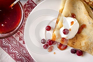 Pancakes with sour cream, berries, cranberries and jam on a plate, tea, jam on a towel with a red pattern on a white