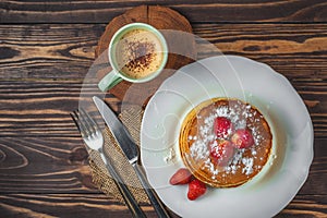 Pancakes with strawberries, icing sugar and a cup of cappuccino for breakfast on a wooden background in a rustic style