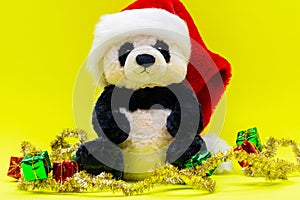 Panda Bear plush toy with Red Christmas Santa Hat surrounded by Tinsel Garlands on bright yellow background. Winter Holidays