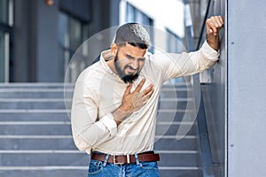 Panic and heart attack on the street. A young Indian man is standing near a building, leaning against a wall and holding