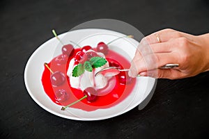 Panna cotta with cherry syrup in white plate and hand with spoon on a dark background and berries. Traditional italian dessert.