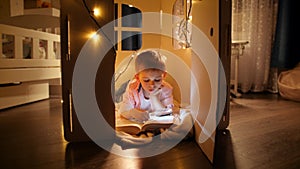 Panning shot of little smart boy reading big book at night with flashlight. Concept of child education and reading in