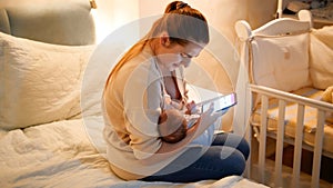 Panning shot of mother using smartphone giving breast to her hungry baby son on bed at night. Concept of healthy and