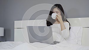 Panning shot of woman drinking a cup of coffee and using laptop computer on bed