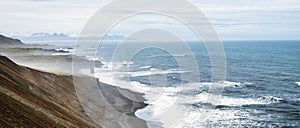 Pano of steep cliffside with volcanic beach and waves crashing to shore.